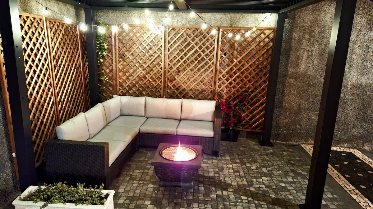 An outdoor seating area features a large, L-shaped sectional sofa arranged around a fire pit. The space is enclosed by decorative wooden lattice panels, with warm lights strung overhead, enhancing the inviting ambiance. Potted plants add a touch of greenery to the corners.