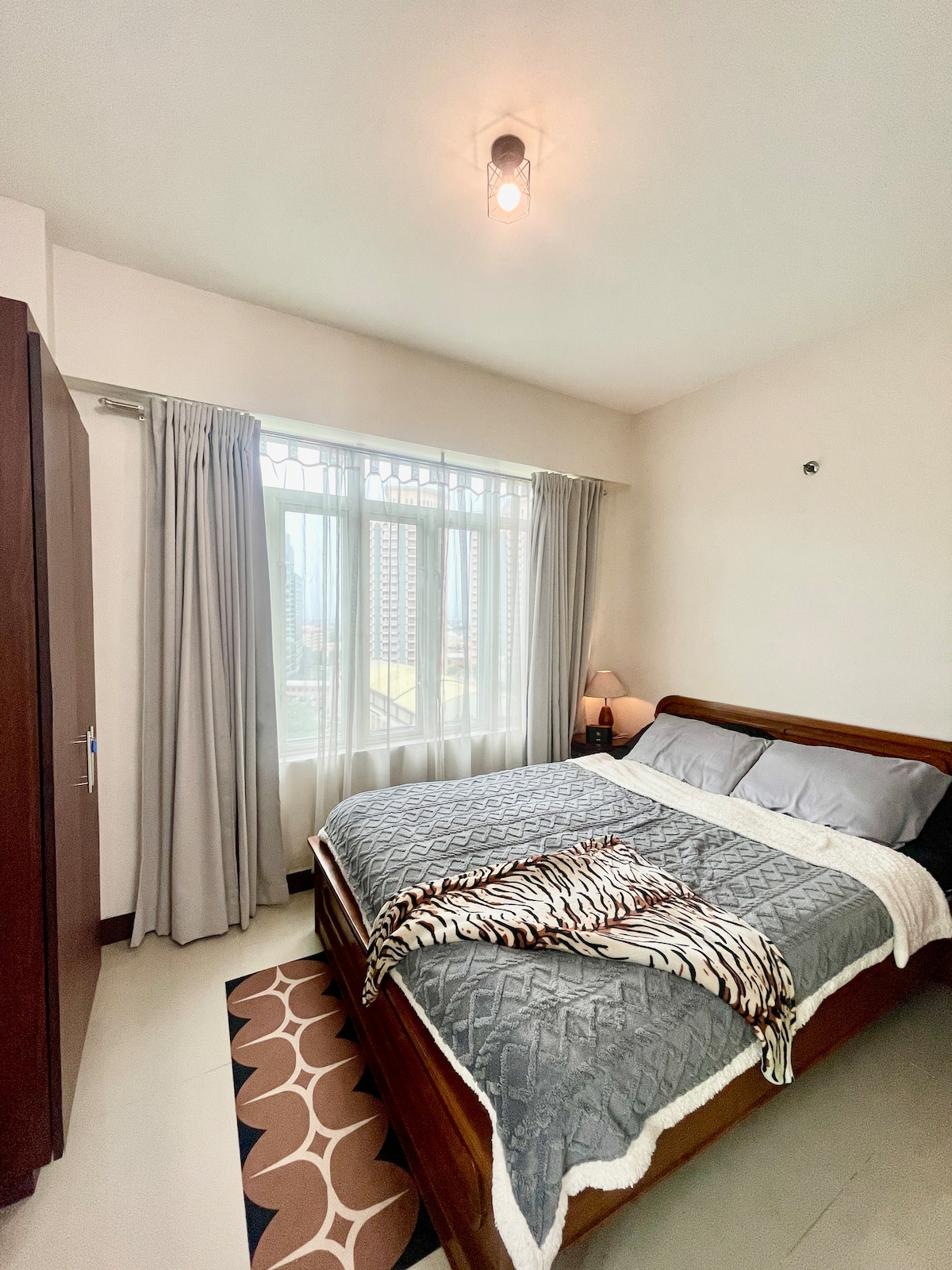 A well-appointed bedroom features a spacious bed with a soft blanket, complemented by textured pillows. Natural light filters through large windows dressed with sheer curtains, while a bedside lamp offers gentle illumination. A wardrobe stands against the wall, enhancing the room's functionality.