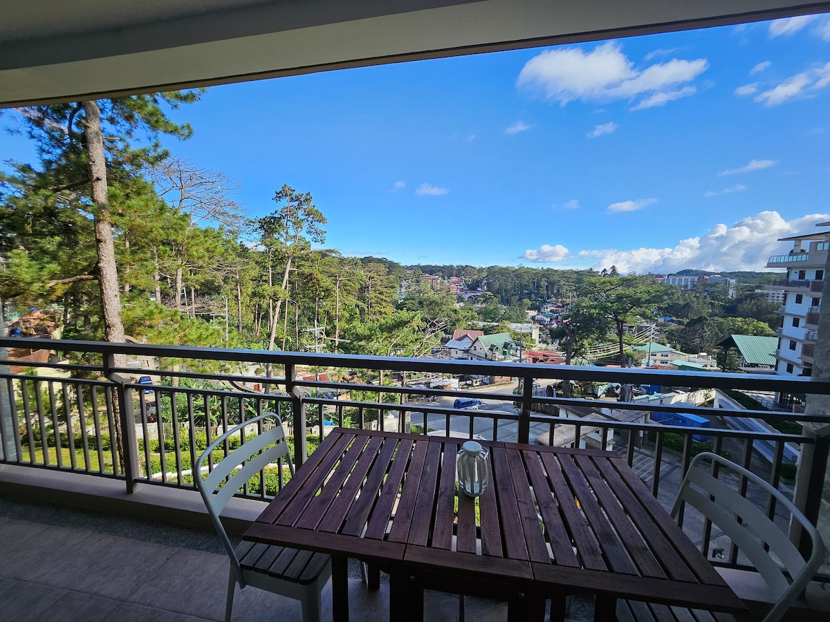 A private balcony features a wooden dining table with chairs, providing an outdoor space to enjoy meals. The view includes a lush green landscape with pine trees and distant buildings, under a clear blue sky.