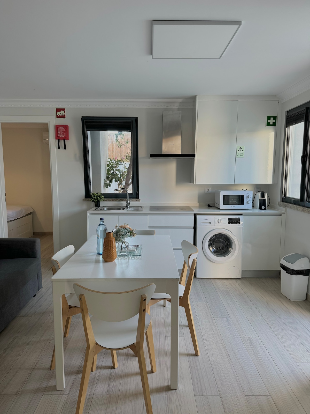 A modern kitchen area is displayed, featuring white cabinetry and stainless steel appliances. A dining table with four chairs occupies the center, while a washing machine is positioned beside the countertop. Natural light enters through two windows, contributing to an airy atmosphere.