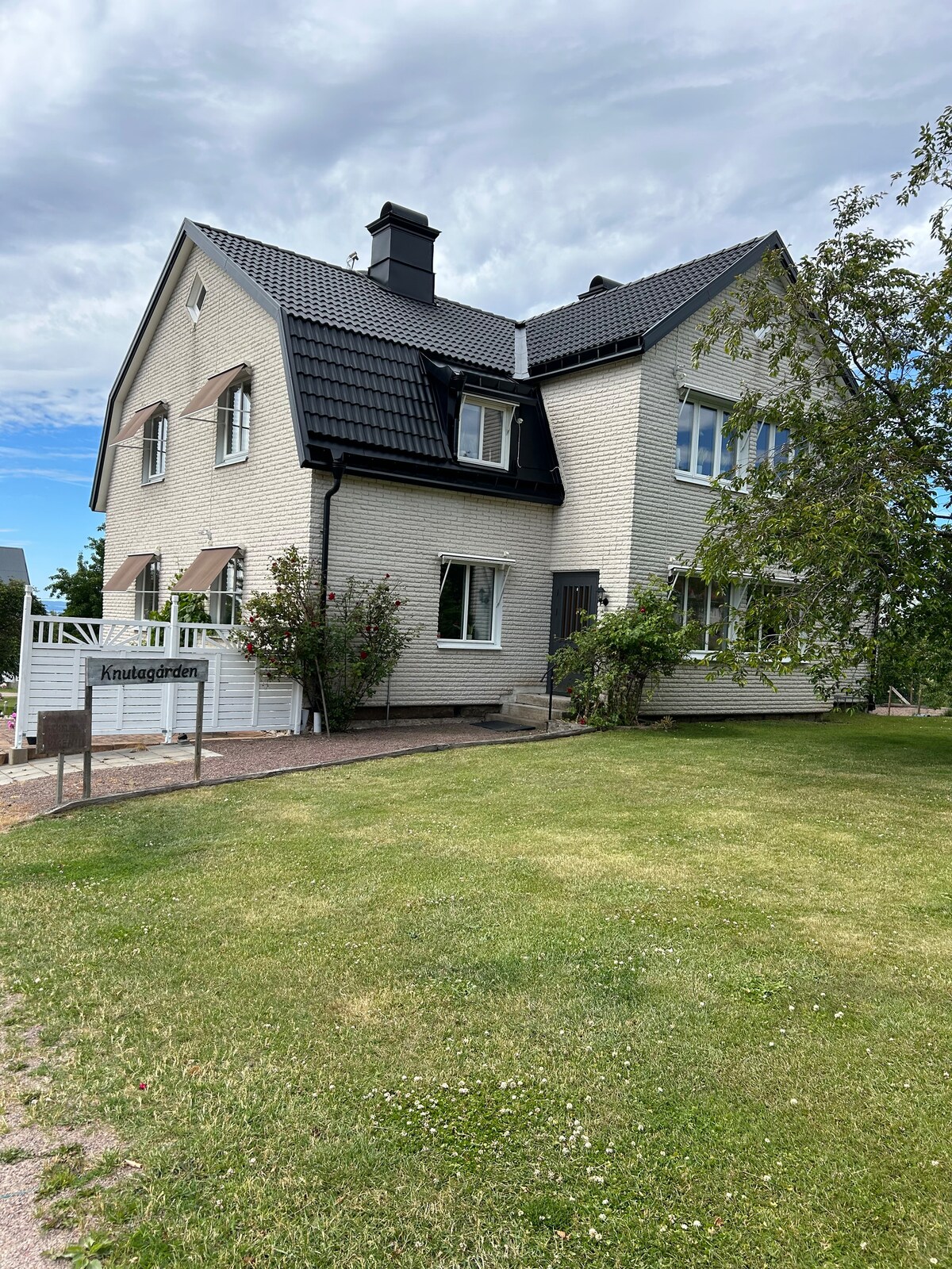 A two-family house is shown with a light-colored exterior. The front yard features well-maintained grass and young trees. A spacious patio area is accessible from the main entrance, and large windows provide ample natural light. The surroundings are quiet and inviting.