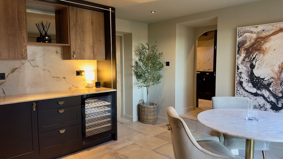 A contemporary kitchen is featured with dark cabinetry and marble accents. A wine cooler is integrated into the cabinetry. A round dining table is positioned nearby, surrounded by elegantly upholstered chairs. Natural light enhances the stone floor and modern decor.