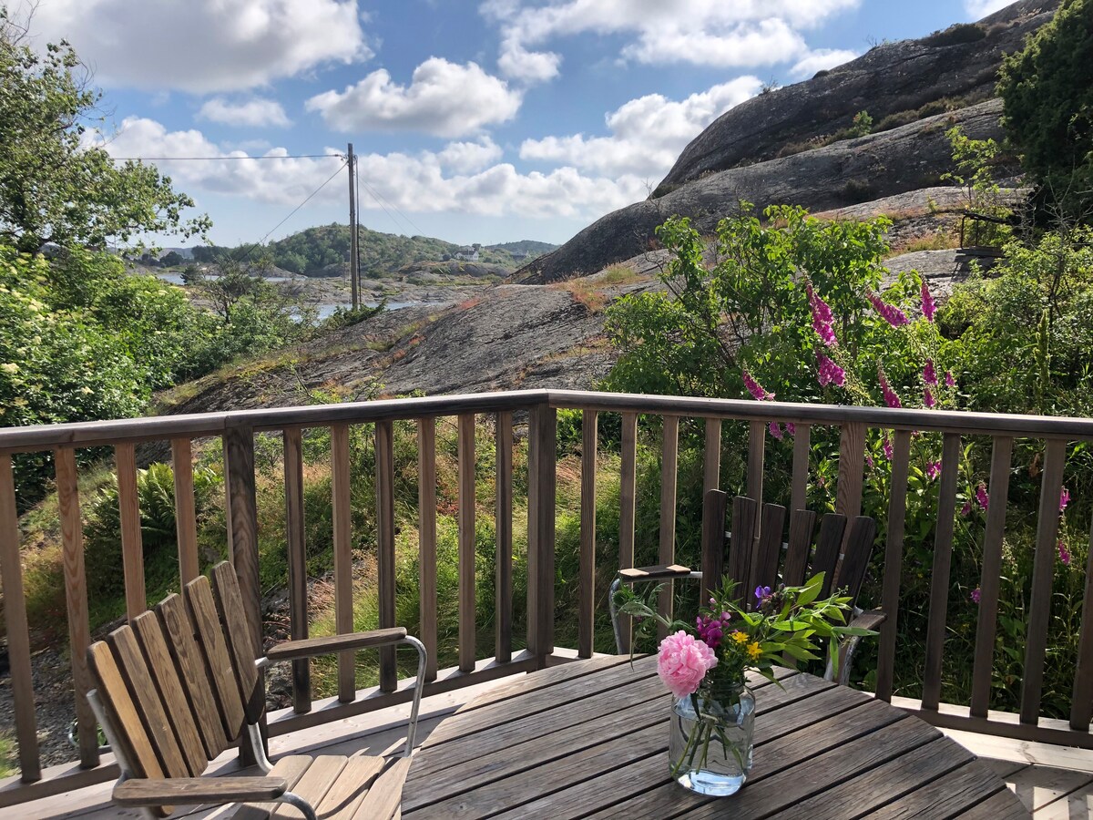 A wooden deck is shown, furnished with two chairs and a small table holding a vase of fresh flowers. Green foliage and blooming plants surround the area, while rocky hills and a cloudy sky provide a natural backdrop.