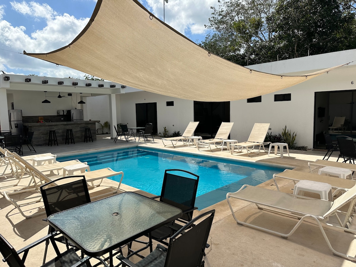 An outdoor area features a refreshing pool, surrounded by lounge chairs and shaded by a large canopy. Several tables and chairs are positioned nearby, offering space for relaxation and dining. The backdrop includes light-colored walls and lush greenery, creating a serene environment.