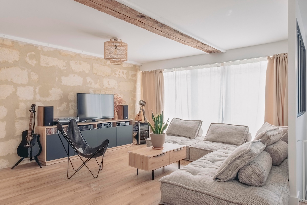 A well-lit living area is furnished with a sectional sofa and a coffee table. A television is mounted on a wall unit, accompanied by a guitar in the corner. Natural light flows through large windows, covered with sheer curtains, enhancing the neutral color palette.