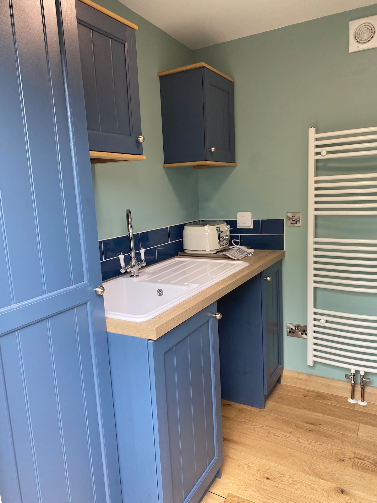 A compact kitchen area is presented, featuring blue cabinetry and wooden countertops. An electric kettle is situated on the counter beside the sink. Light-colored wooden flooring complements the room's design, while a heated towel rail is mounted on the wall, enhancing functionality.