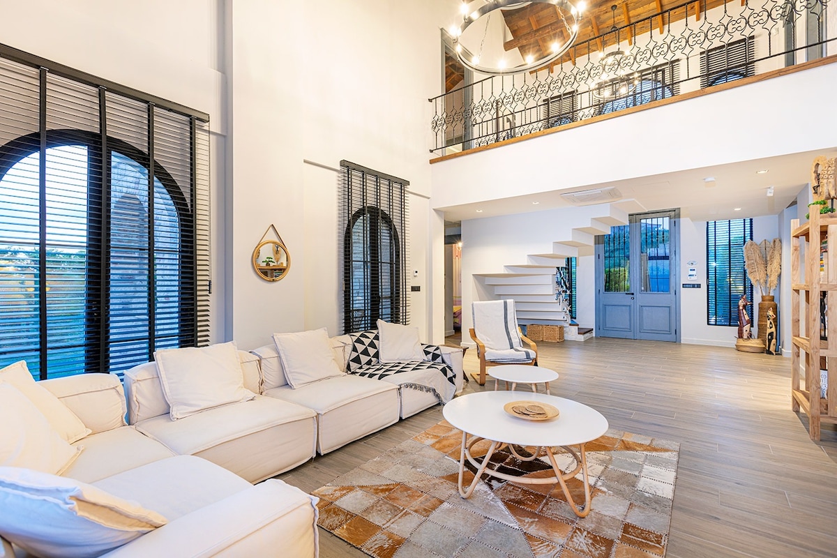 A bright and airy living room features high ceilings and large arched windows, allowing natural light to flood the space. A comfortable sectional sofa is arranged around a round coffee table, while a decorative area rug adds a touch of warmth. A modern staircase leads to the upper level.
