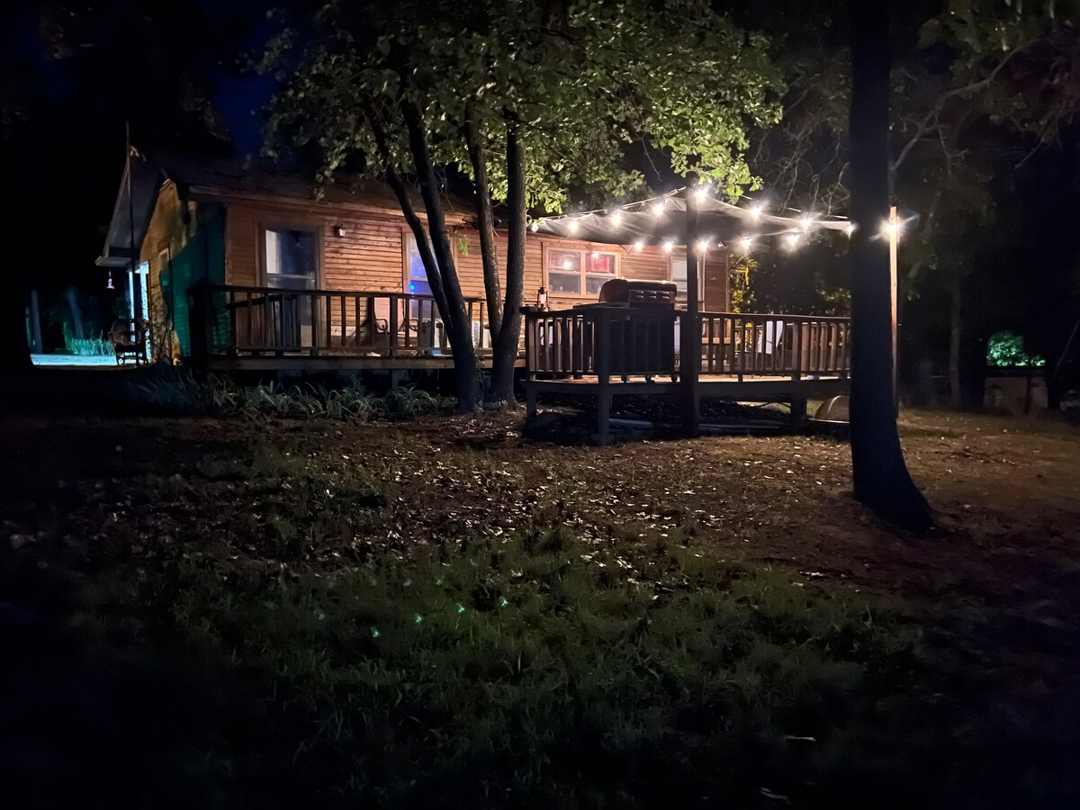 The cabin is depicted at night, illuminated by soft string lights over the spacious deck. Surrounding trees create a natural boundary, while the wooden exterior reflects a warm glow from the home, offering an inviting atmosphere in a wilderness setting.
