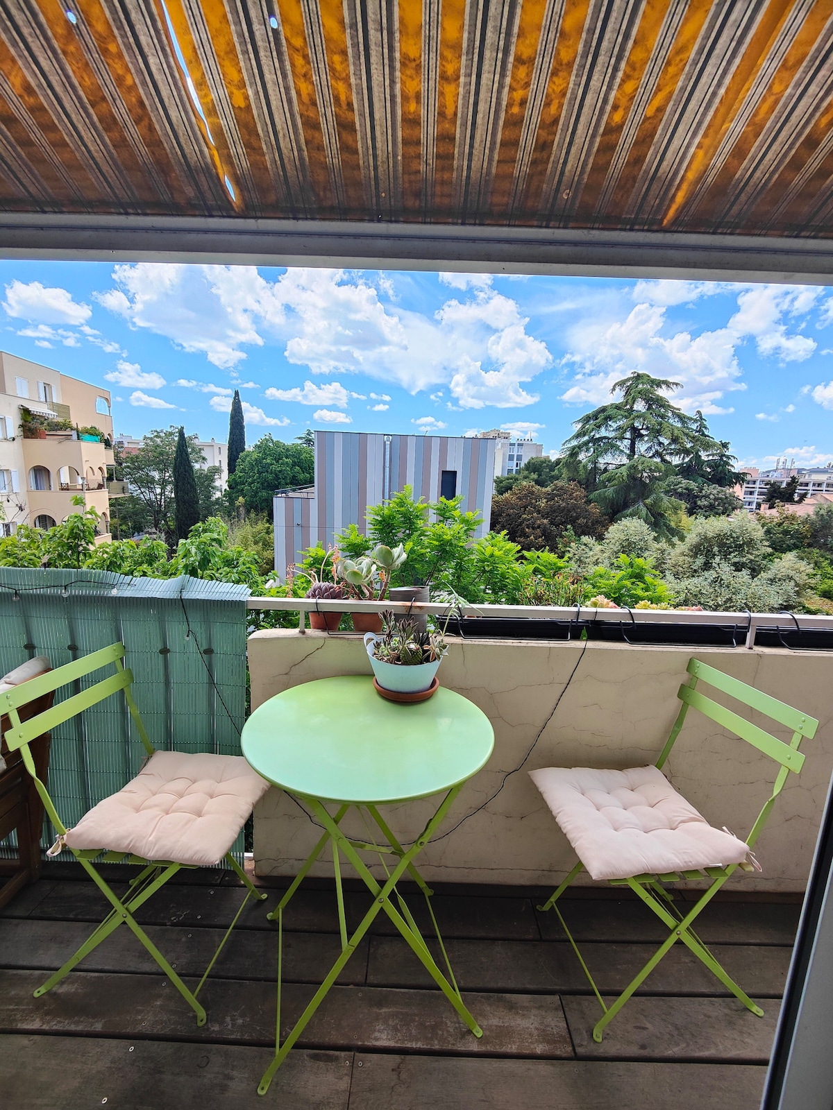 A small balcony features a round green table with two matching chairs, each adorned with soft cushions. Lush greenery is visible in the surrounding area, along with a bright sky filled with scattered clouds creating a serene outdoor space.