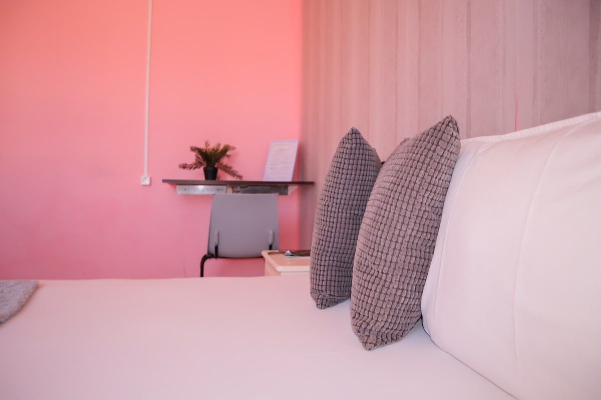 A cozy sleeping area features a spacious bed adorned with patterned gray pillows. Soft light enhances the warm pink wall backdrop. A small desk is positioned nearby, complemented by a potted plant, creating a functional yet inviting space.