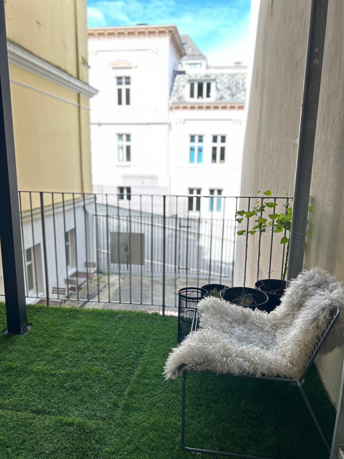 A small balcony is visible, featuring artificial grass flooring and a minimalist chair covered with a soft, white throw. Potted plants are arranged on the floor near the chair. The view includes neighboring buildings with various window styles and architectural details.