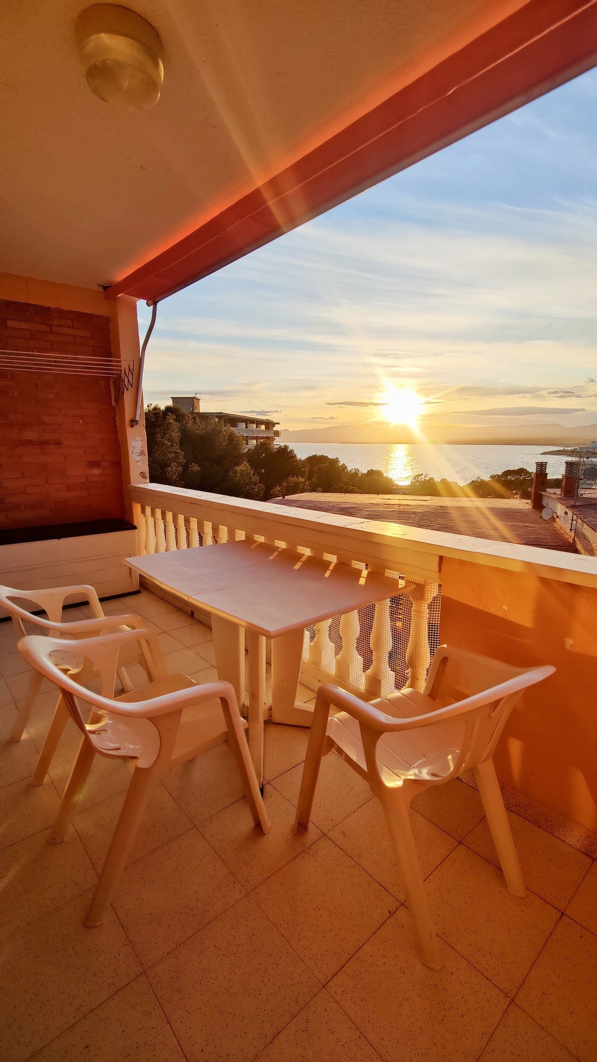 A balcony is observed, featuring a small table and two white chairs. The view showcases a sunset over the sea, with vibrant colors illuminating the horizon. The surrounding area includes trees and buildings, enhancing the tranquil atmosphere.