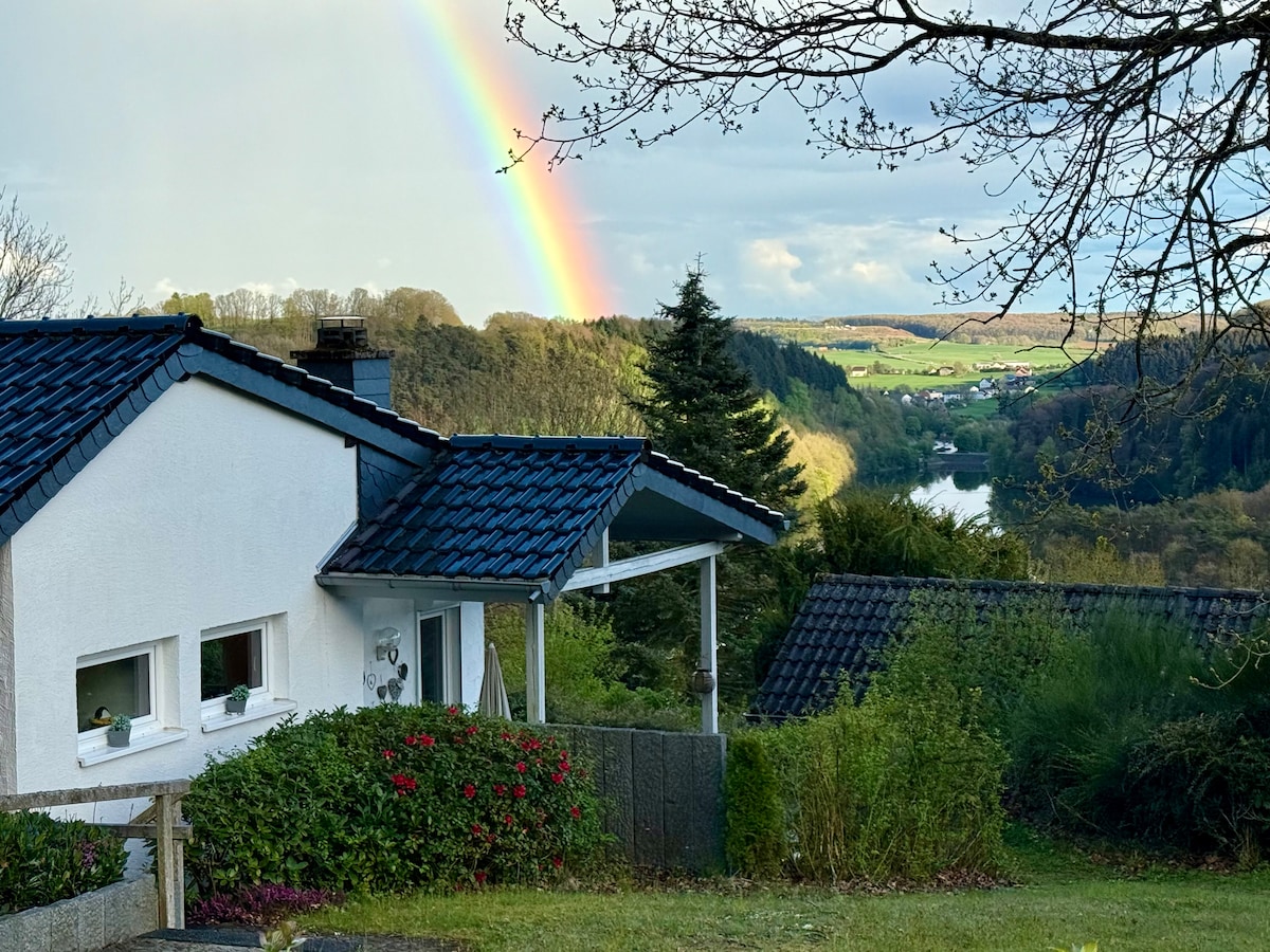 A view of the holiday home is presented, showcasing a sloped roof and windows facing a vibrant landscape. A rainbow arcs over the greenery and rolling hills in the background, highlighting the natural beauty of the surroundings.