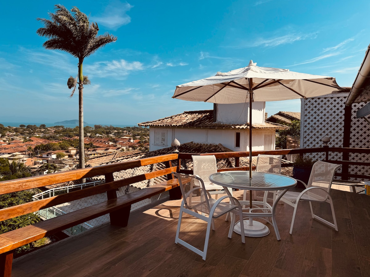 A spacious outdoor terrace is presented, featuring a round glass table surrounded by four white chairs. A large umbrella provides shade, while a palm tree adds greenery. The background reveals a coastal view and charming rooftops under a clear blue sky.