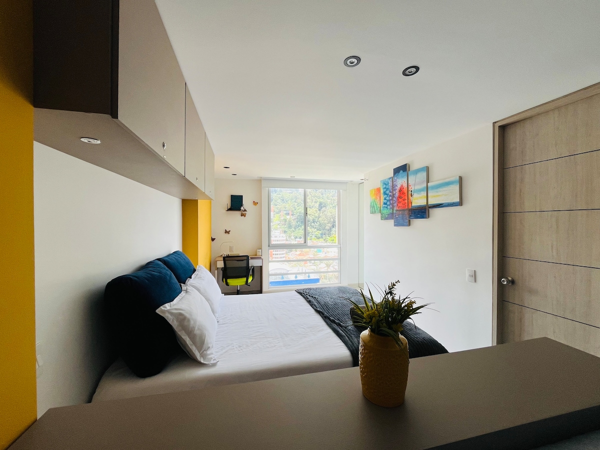 A studio apartment is presented featuring a spacious bed with crisp white linens and a dark throw blanket. Bright yellow walls enhance the setting, while a small desk and a potted plant add functionality. Large windows showcase views of the surrounding mountains and natural light.