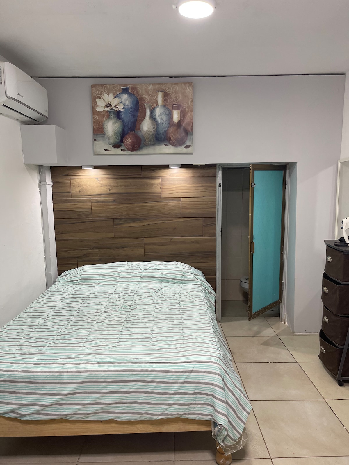 A small room features a wooden bed with a light striped blanket, complemented by a wall-mounted art piece depicting vases. An adjacent closet with a door leads to a private bathroom, while the room is illuminated by ceiling lights and includes neutral-toned tiles.