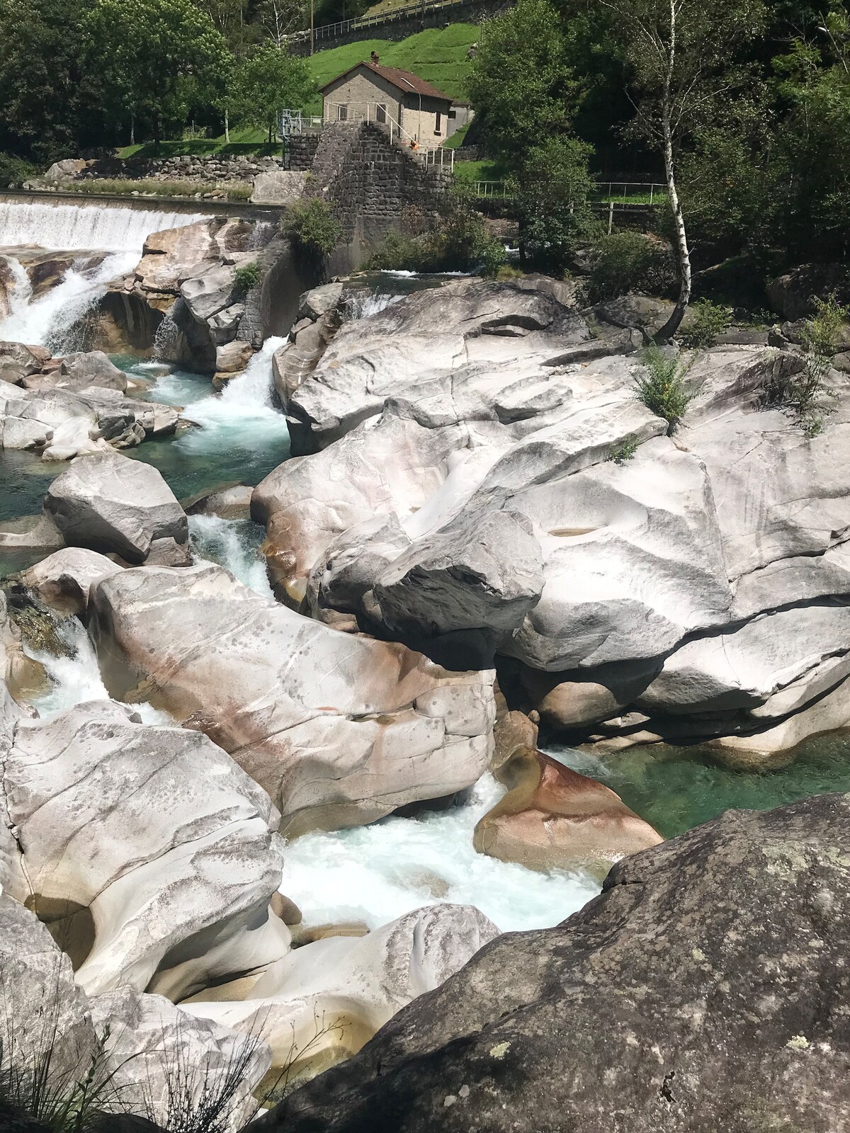 A serene landscape showcases a flowing river surrounded by smooth, large stones. The water cascades over rocky formations, creating a gentle current. Lush greenery is visible in the background, along with a stone building partially obscured by trees.