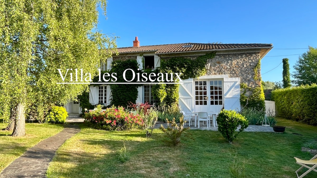 A charming stone house is framed by lush greenery and vibrant flowers. The exterior features white shutters and a welcoming entryway, with a well-maintained garden set against a clear blue sky.