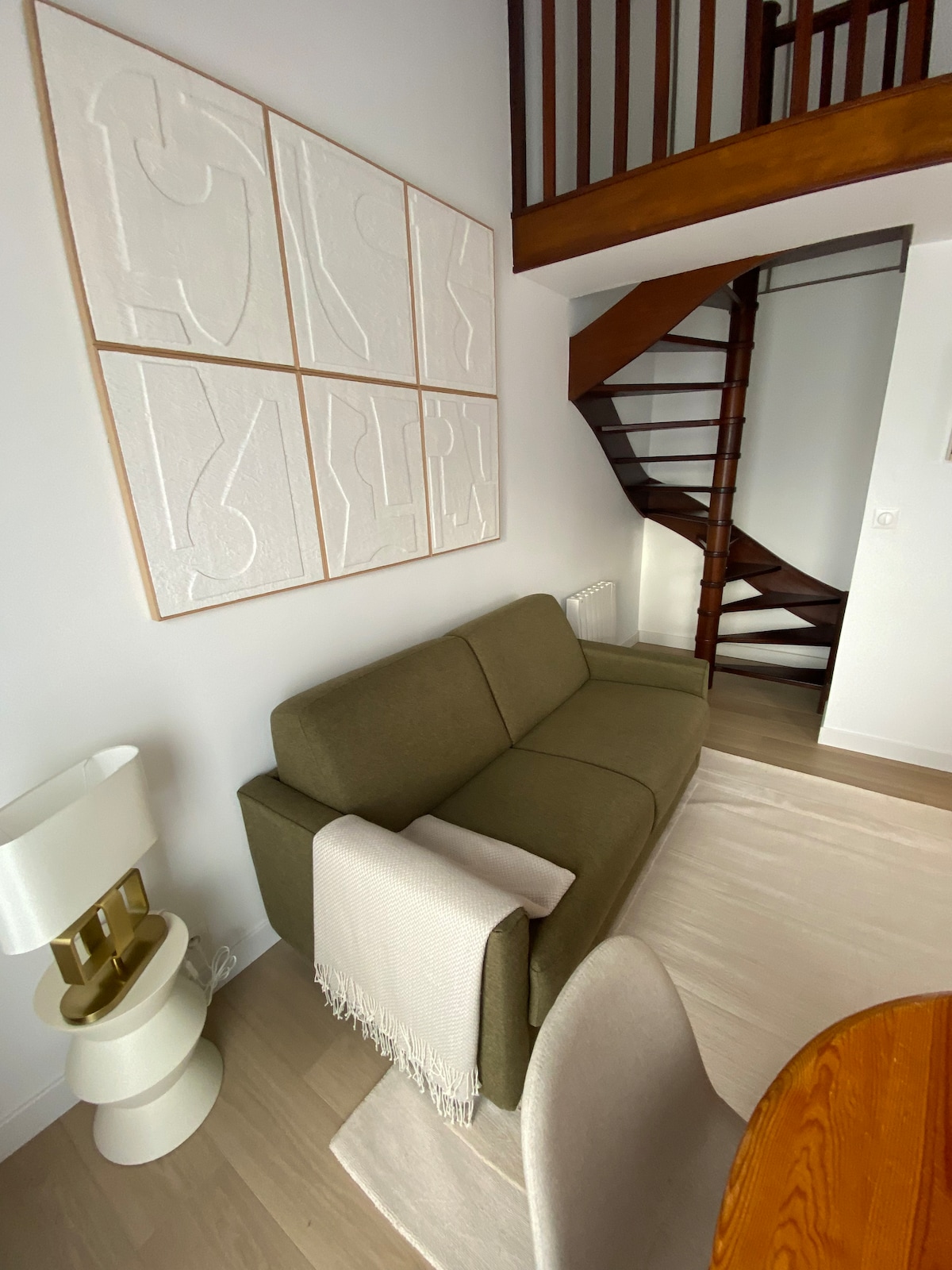 A comfortable sofa, draped with a light-colored throw, sits in the living area, accompanied by a stylish lamp. A round dining table is nearby, and a spiral staircase ascends to the upper level, adding architectural interest to the bright space.