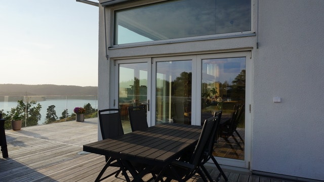 A spacious terrace features a wooden dining table surrounded by black folding chairs. Large glass doors provide views of the serene landscape, reflecting the natural light and enhancing the connection to the outdoors.