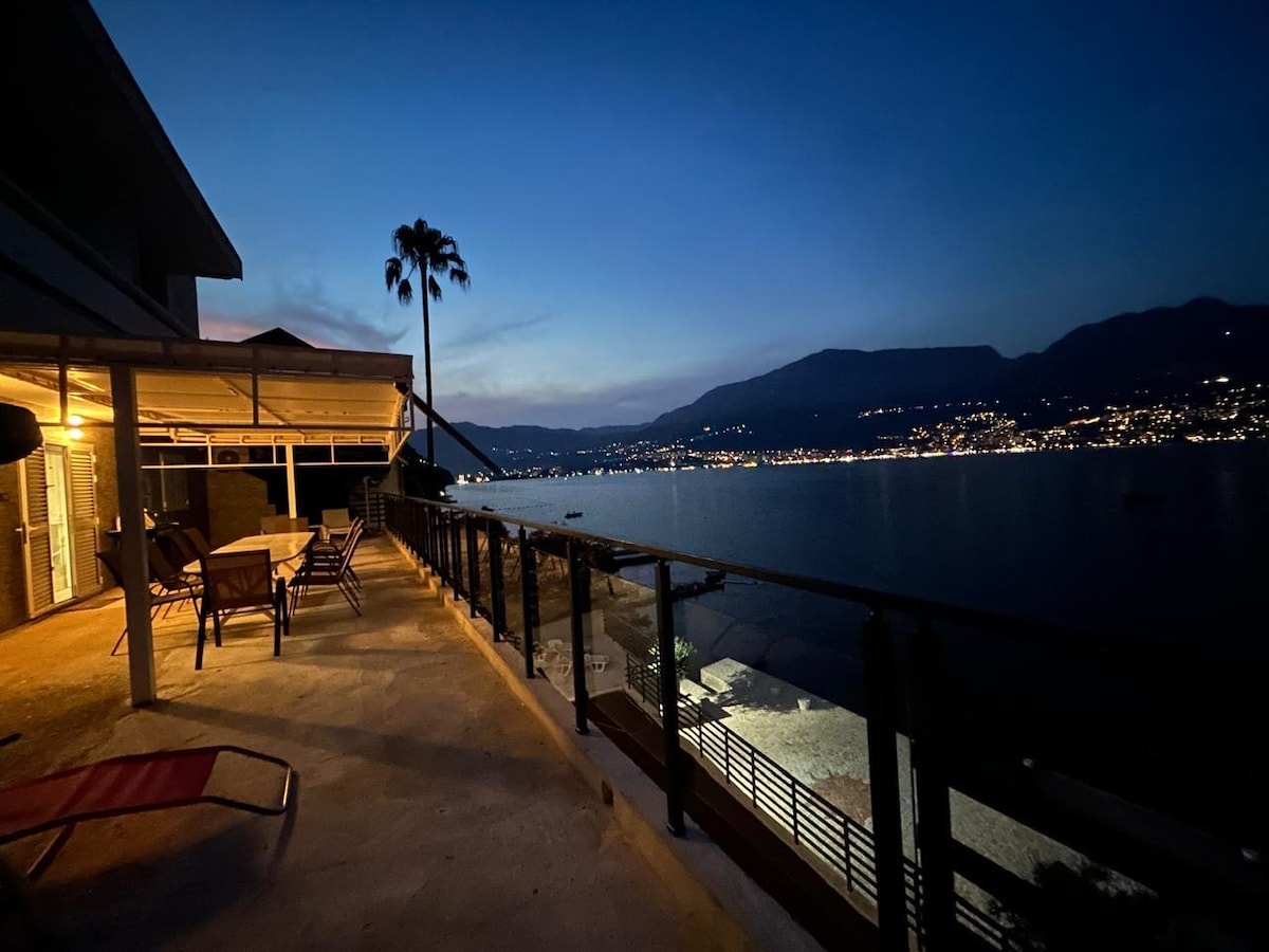 A spacious terrace is illuminated by dusk, featuring an outdoor dining table surrounded by chairs. The view showcases the tranquil sea and twinkling lights of the nearby coastline. A single palm tree is visible, enhancing the serene atmosphere.