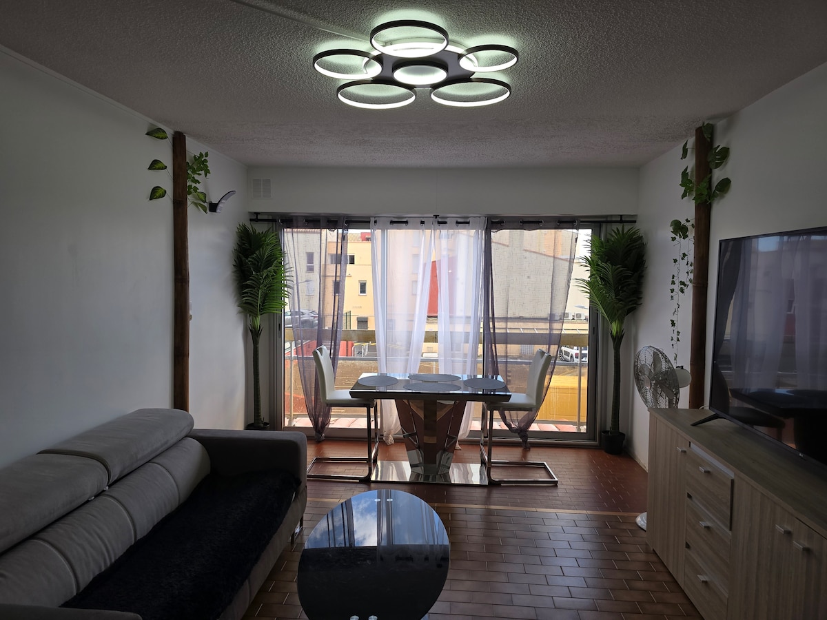 The living area features a modern couch facing a large window. A glass dining table is positioned in front, surrounded by chairs. Soft lighting is provided by a unique ceiling fixture, while indoor plants offer a touch of greenery. Light curtains filter the sunlight.
