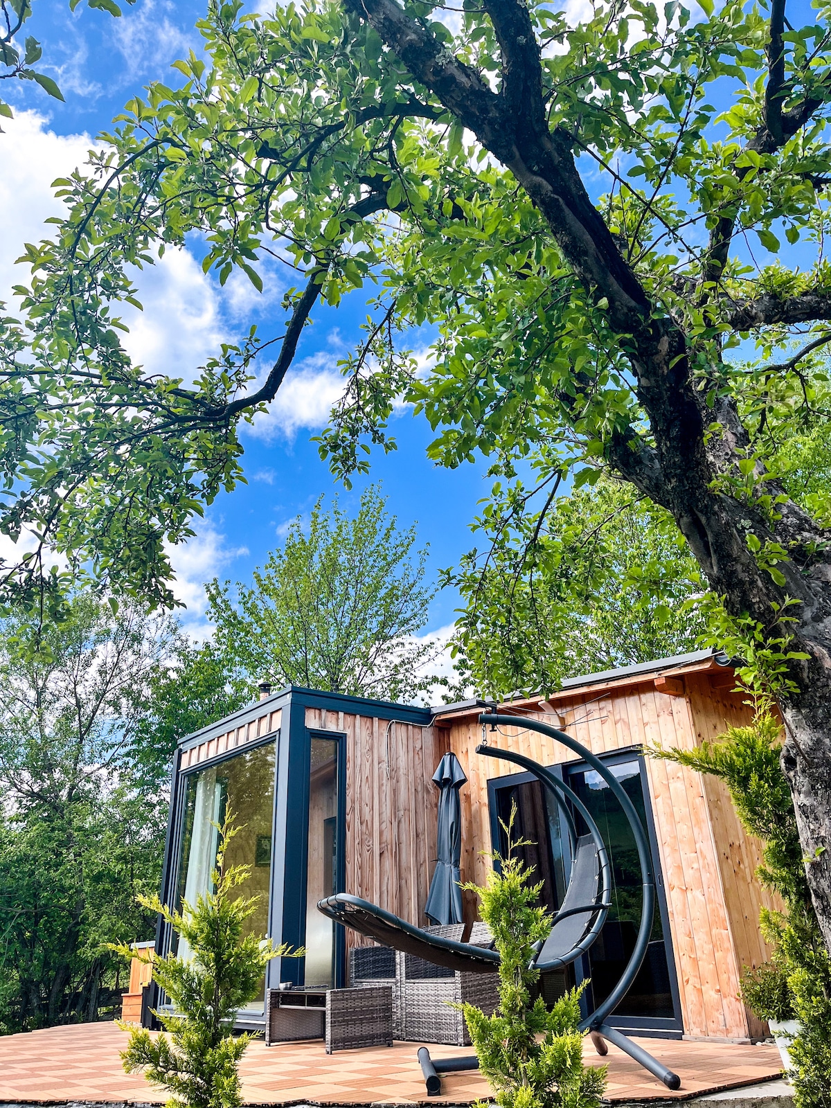 The tiny house showcases large glass doors framed by natural wood siding and greenery, offering a glimpse of the outdoor seating area. Sunlight filters through leaves above, creating a serene outdoor setting for relaxation.