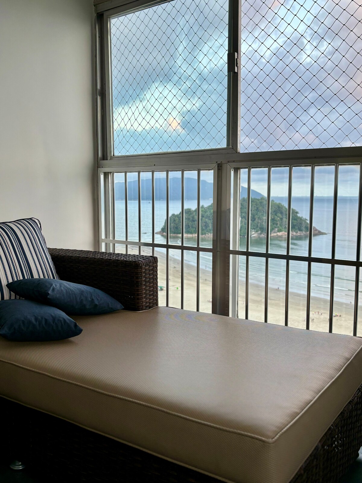 A comfortable seating area features a cushioned lounge with striped pillows, positioned near a large window. The view reveals the coastline with gentle waves and an island in the background, framed by a protective metal grid.