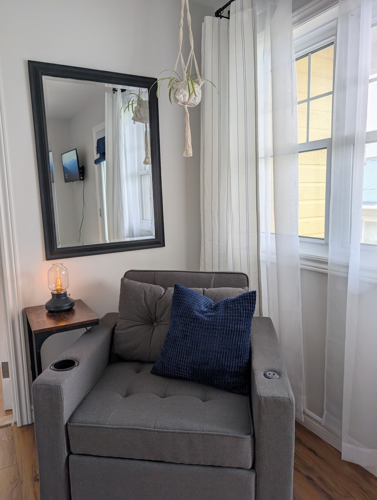 A comfortable gray chair is positioned near a window, with a blue decorative pillow resting on it. A mirror hangs on the wall opposite, reflecting the light flowing in through sheer white curtains. An accent table with a lantern complements the cozy corner.