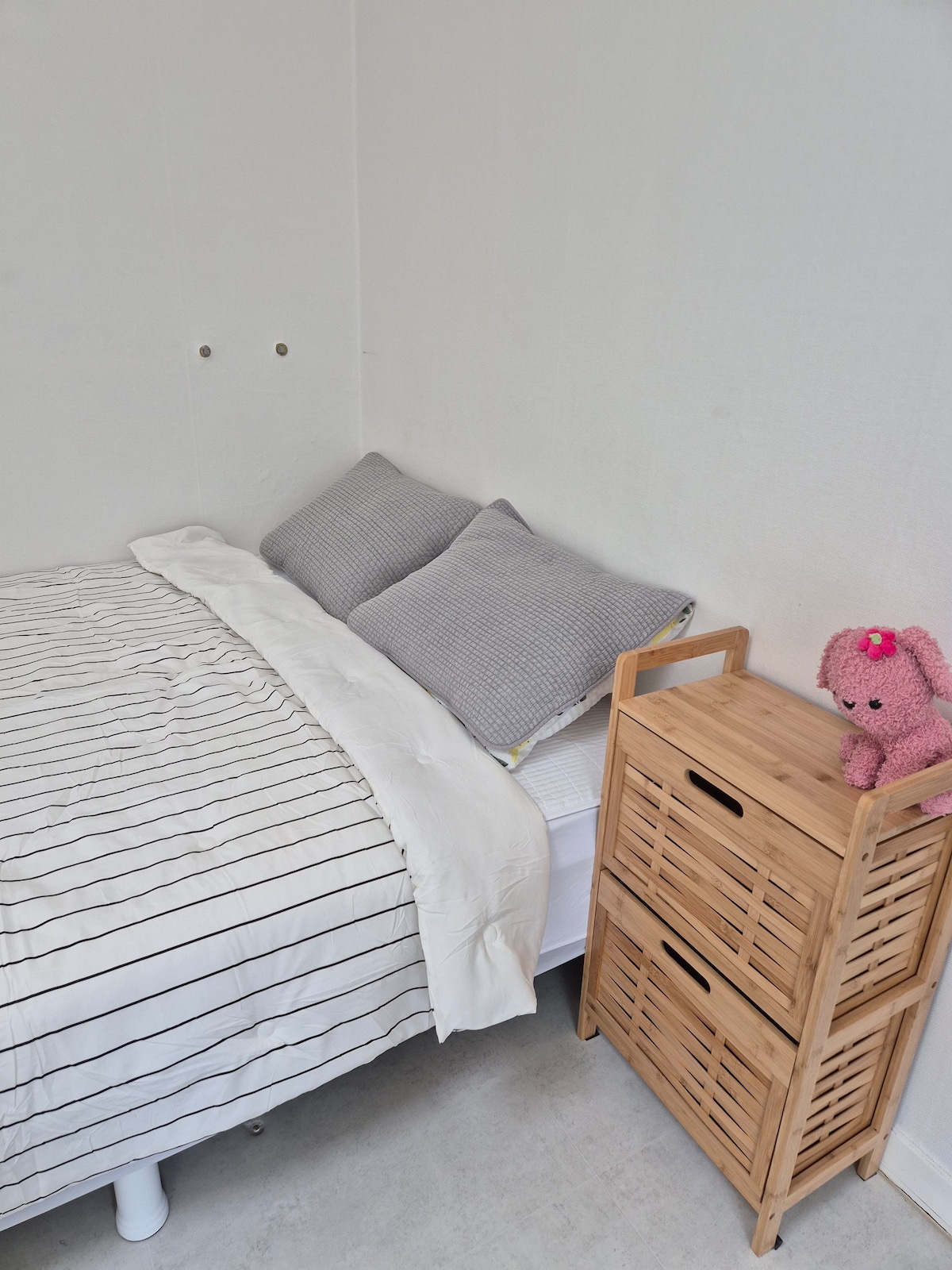 A cozy sleeping area features a spacious bed with a striped blanket and two soft grey pillows. A wooden nightstand with a small handle sits beside the bed, accented by a plush pink toy. The room is brightened by light walls, creating a simple, inviting space.