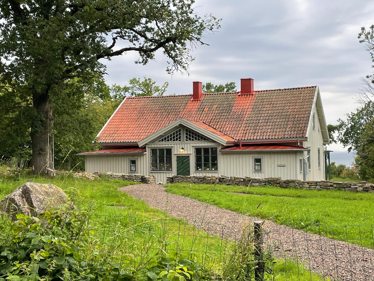 The house features a traditional design with a gabled roof, red accents, and a welcoming entrance. A gravel path leads through lush greenery, framed by natural stone walls. Tall trees are visible in the surrounding area, under a cloudy sky.