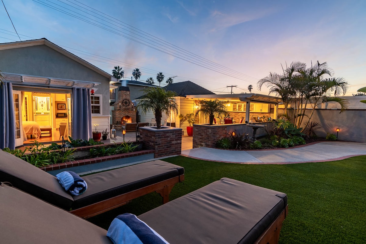 The outdoor space features comfortable lounge chairs positioned on lush green grass, inviting relaxation at dusk. A charming patio area is visible, leading to a warm glow from the kitchen, while a brick outdoor fireplace adds a cozy touch to the serene atmosphere.