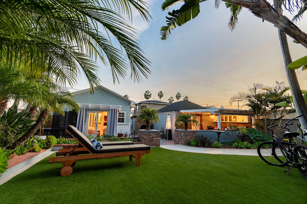 An inviting outdoor space is highlighted, featuring a sun lounger on well-maintained artificial grass. Lush tropical plants and palm trees frame the area. A charming patio with cozy seating and a barbecue area is visible in the background, providing a serene atmosphere for relaxation.