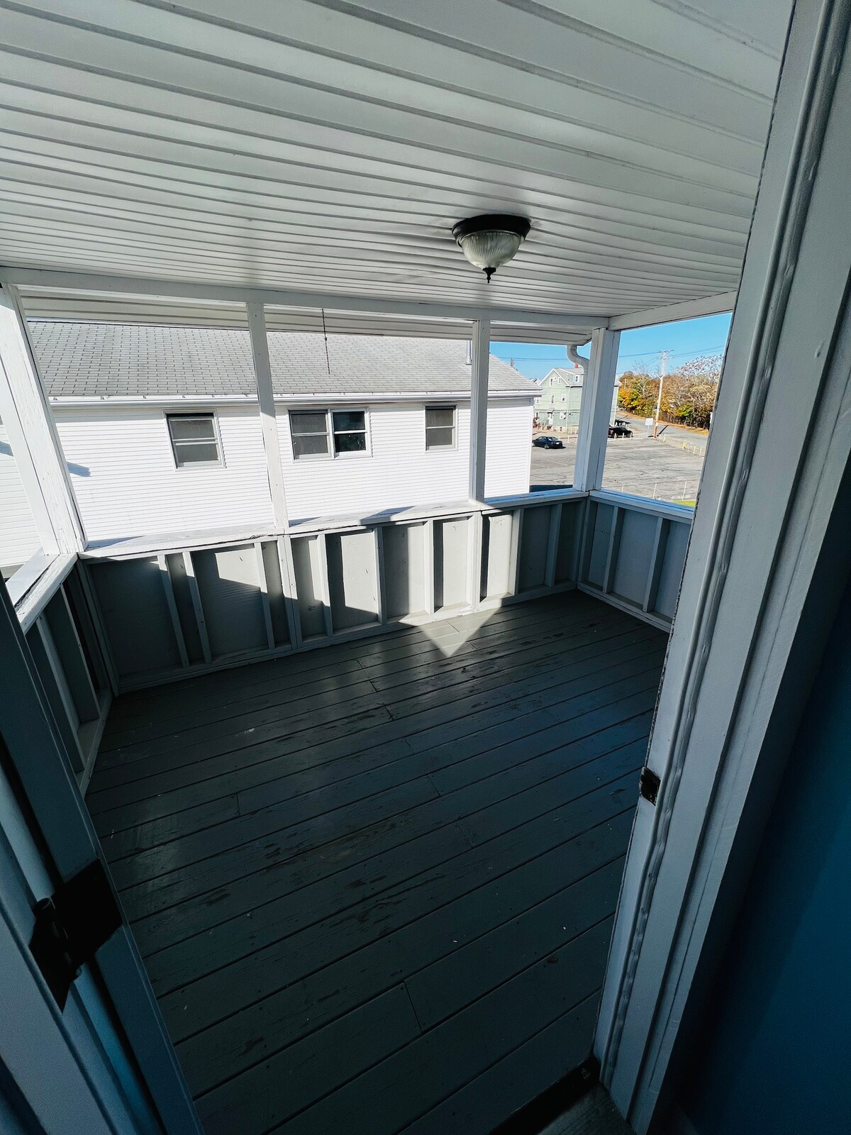Bright and spacious balcony area is revealed, featuring light grey wooden flooring and walls. Natural light seeps in through the open railing, illuminating the space. A light fixture hangs from the ceiling, offering an inviting setting for relaxation.