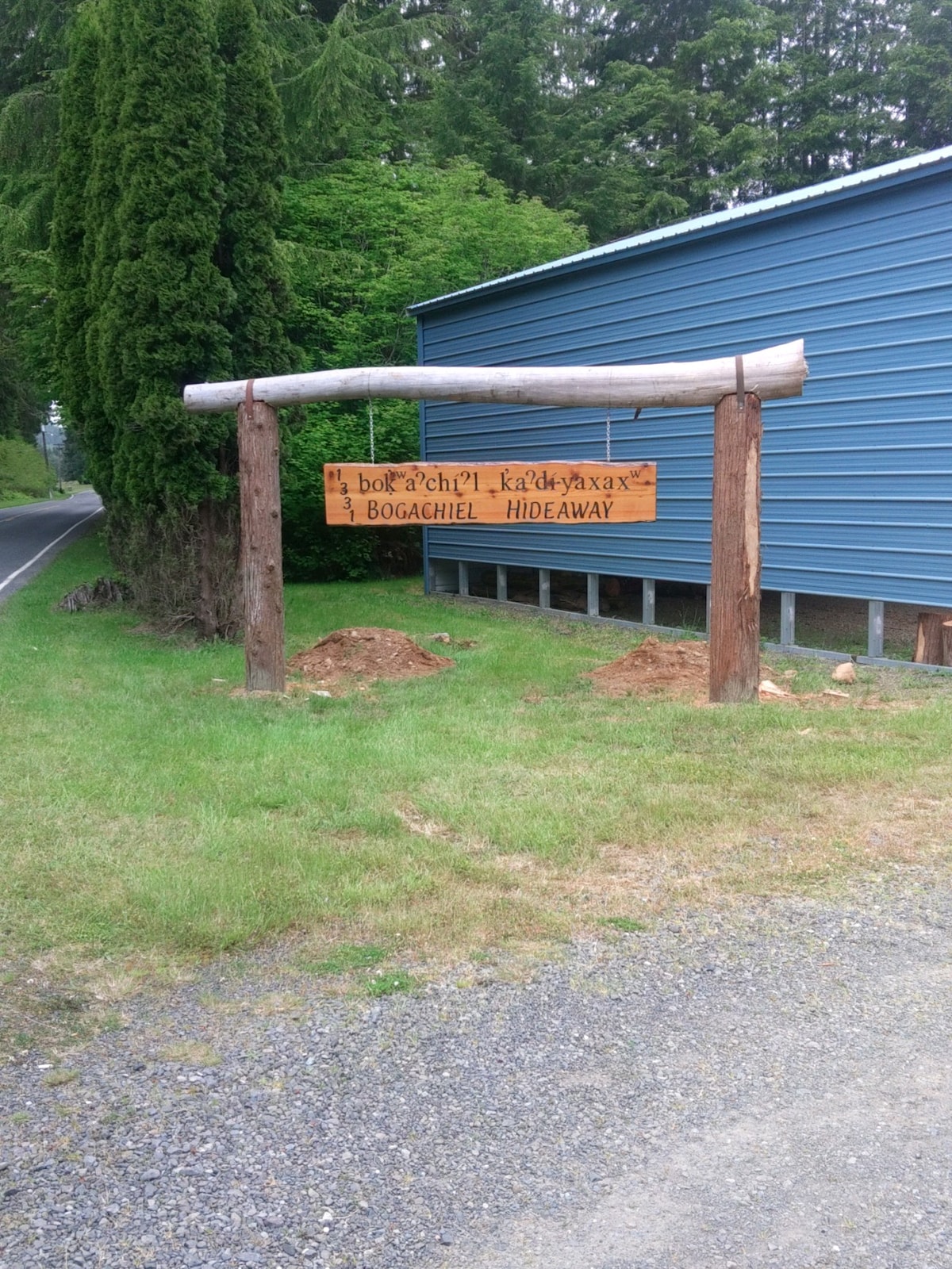 A wooden sign marking the entrance to Bogachiel Hideaway is displayed prominently. The sign, featuring the name in both English and a local language, is supported by a rustic log frame. Lush greenery and gravel lining the roadside are visible in the background.