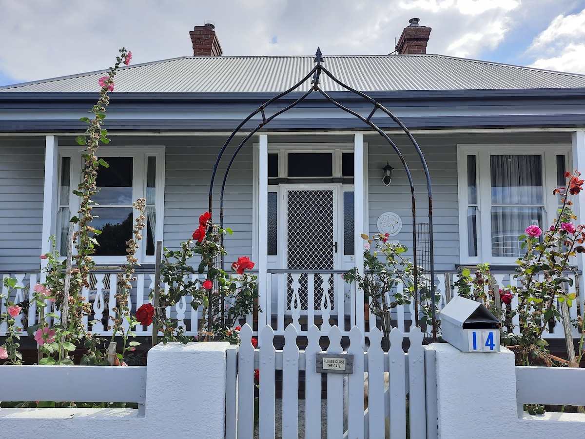 The exterior of a charming cottage features a welcoming front garden adorned with blooming roses. A white picket fence encloses the property, with an archway leading to the entrance. The grey façade is complemented by white window frames and a central door, contributing to its quaint appeal.