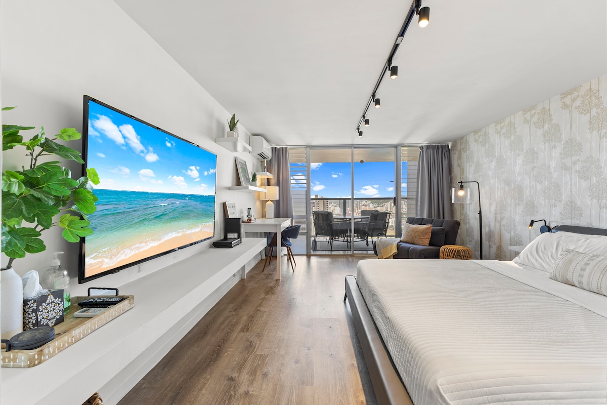 A spacious studio is highlighted by a large 65” Smart TV and a king-size bed featuring fresh linens. Natural light pours in from the sliding glass doors, leading to a balcony. A dedicated work area with a desk and chair complements the modern decor.