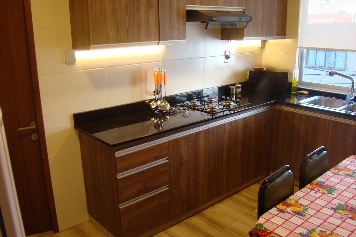 A modern kitchen features wooden cabinetry with a sleek black countertop. A gas stove is positioned beneath a minimalist range hood, while a sink and an array of utensils are visible. A vibrant tablecloth adorns the dining table, adding color to the space.