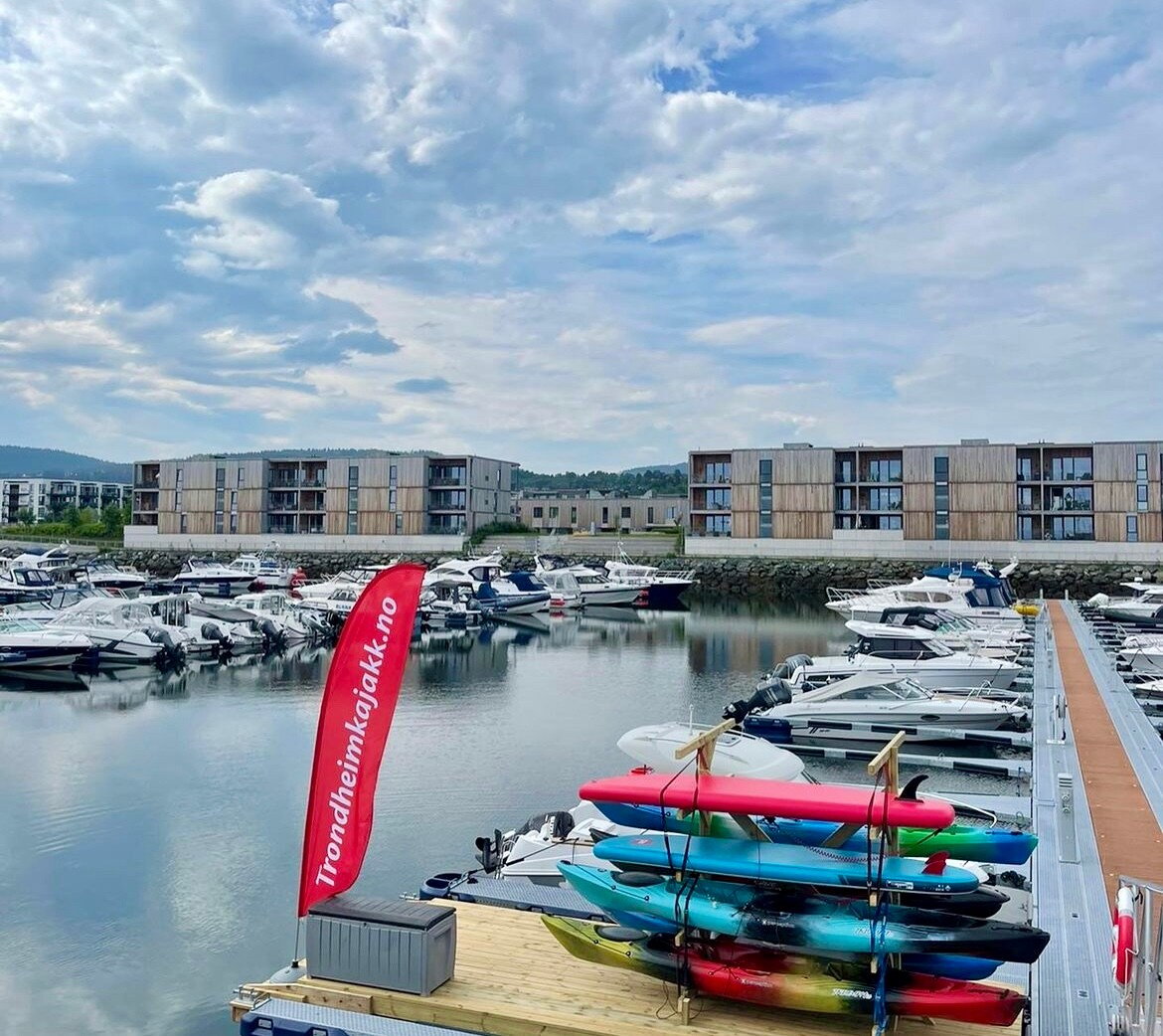 A marina scene is displayed, featuring a calm waterway lined with boats of various sizes. Colorful kayaks are arranged neatly on a dock, while modern waterfront buildings are visible in the background under a partly cloudy sky.