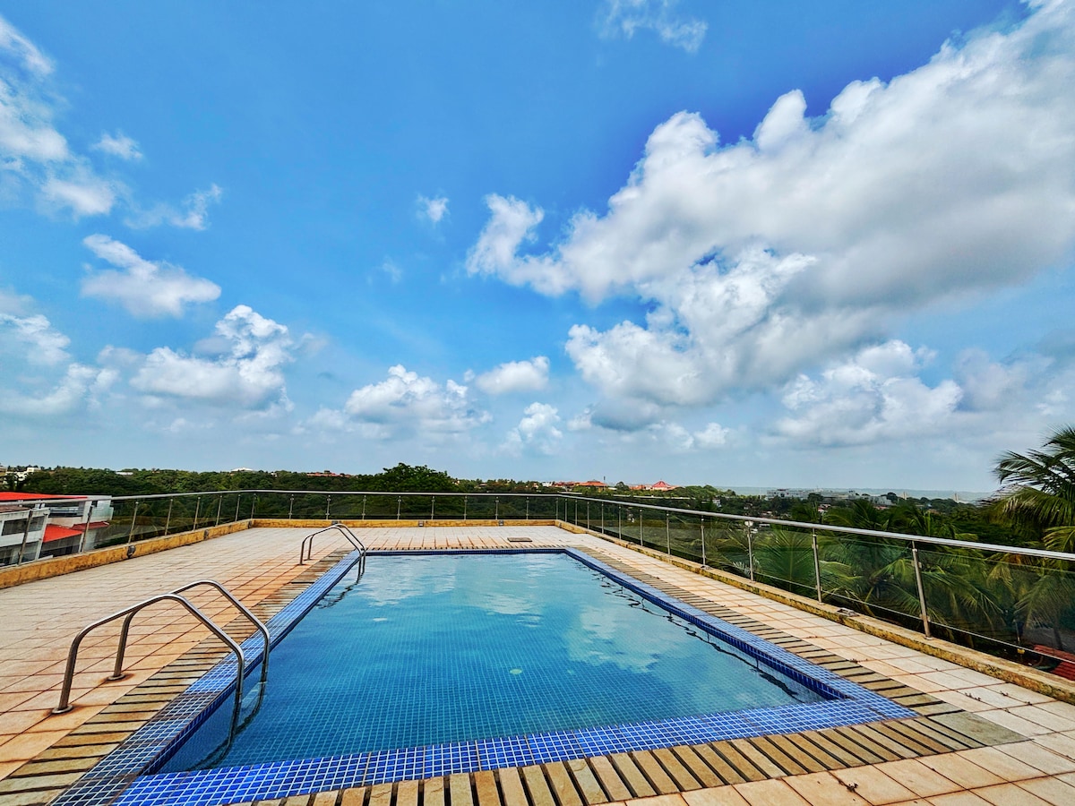 A rooftop swimming pool is featured, surrounded by a spacious deck. Clear blue water reflects the sky, which is dotted with fluffy clouds. The pool's edges are lined with tiles, and a set of stairs leads into the water, offering an inviting view of the valley below.