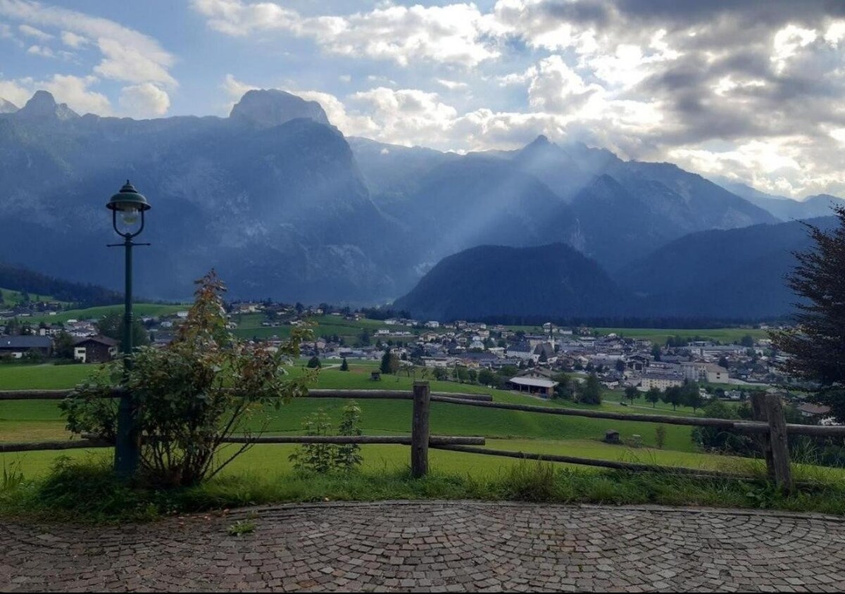A scenic view showcases rolling green fields and a small village nestled among mountains. Sunlight breaks through the clouds, casting rays over the landscape. A stone pathway and wooden fence frame the scene, enhancing the natural beauty of the surroundings.