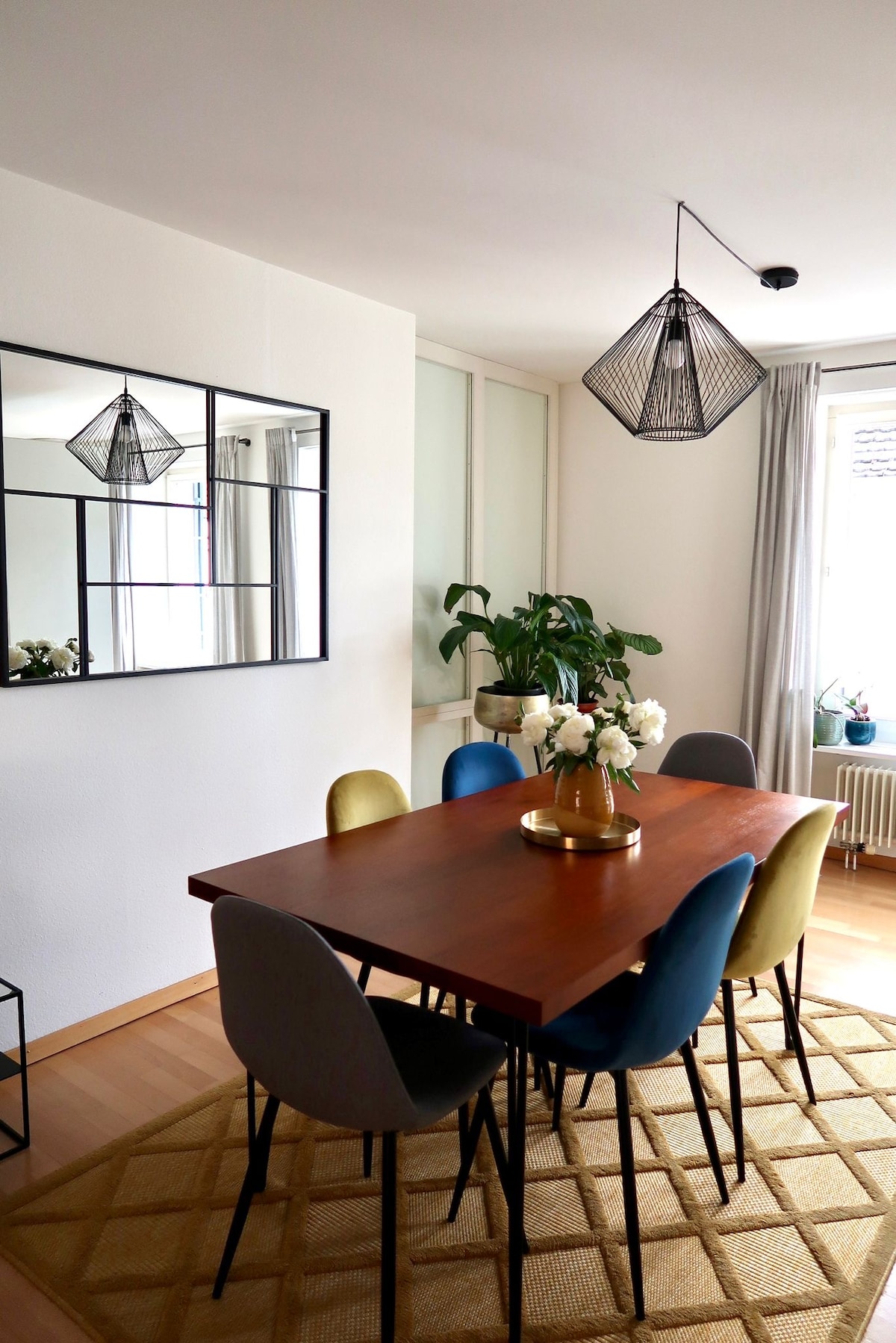 A dining area is presented with a wooden table surrounded by six upholstered chairs in varying colors. A modern pendant lamp hangs above, illuminating a vase of flowers at the center. A decorative mirror and green plants add subtle touches against the light walls.