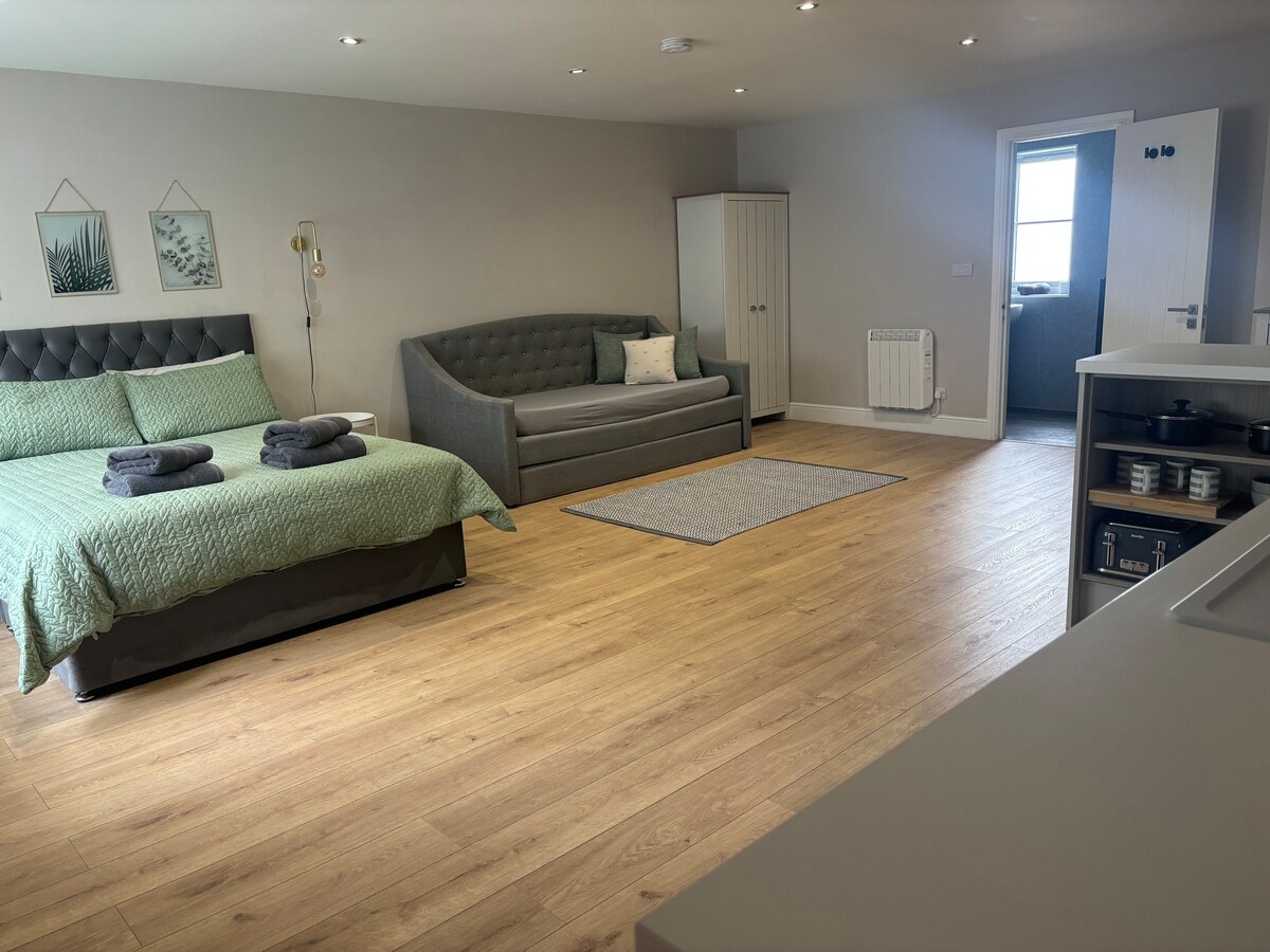 The spacious living area includes a king-size bed with a green blanket and two neatly arranged towels on top. A day bed with a neutral cushion sits adjacent to the bed. Light-colored wood flooring adds warmth, while a door leads to an additional room.