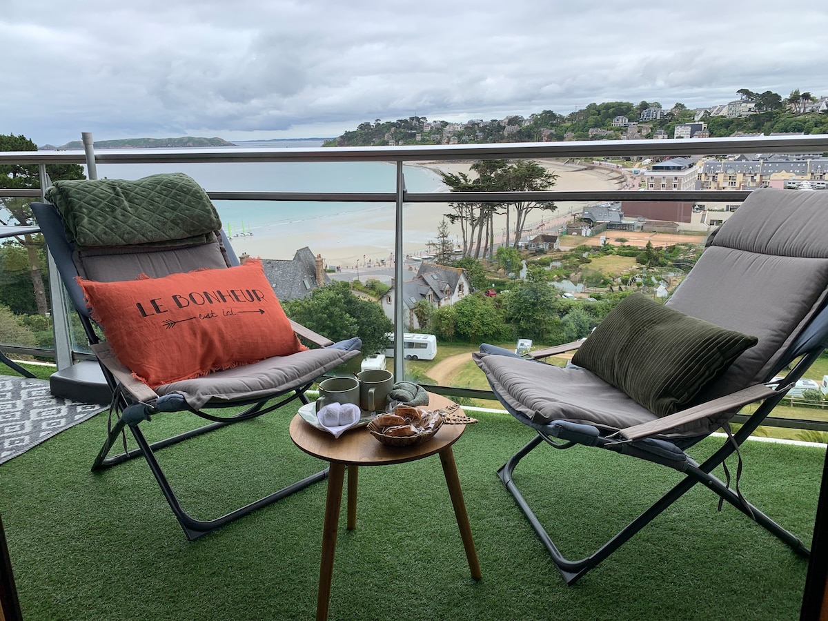 A terrace features two comfortable loungers facing a scenic view of the beach and coastline. A small table is set between them, enhancing the inviting atmosphere. Greenery fills the space, and coastal charm is apparent in the surroundings.