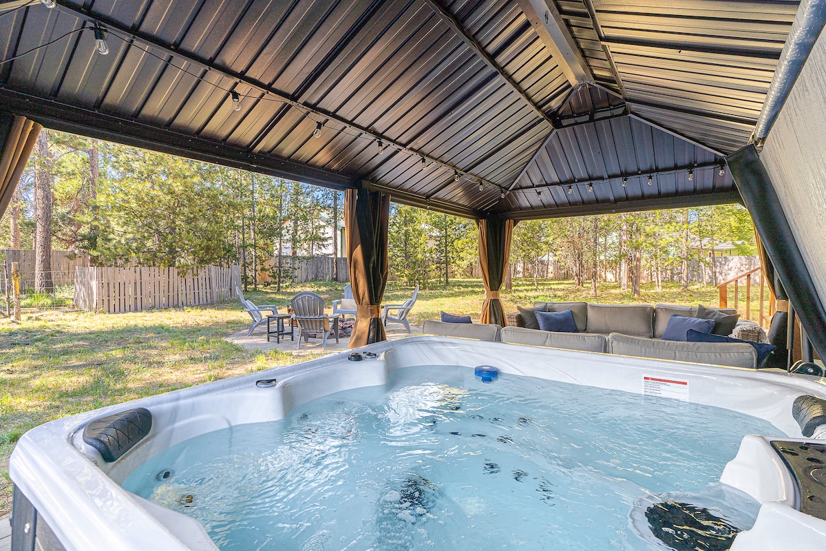 A spacious outdoor gazebo shelters a hot tub, with clear water inviting relaxation. Surrounding seating areas are arranged for comfort, complemented by lush greenery in the backyard. Soft, natural light filters through the open space, enhancing the tranquil setting.