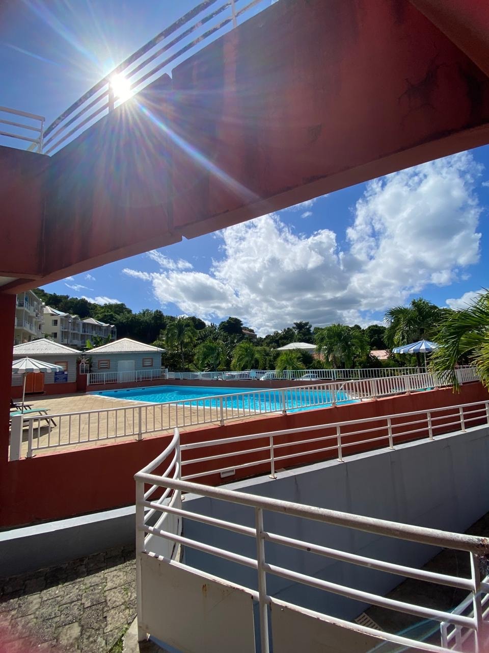 A sparkling swimming pool is visible through a structural archway, surrounded by lounge chairs and lush greenery. Bright blue skies with scattered clouds create a vibrant backdrop, while buildings are seen in the distance, contributing to the inviting atmosphere of the area.