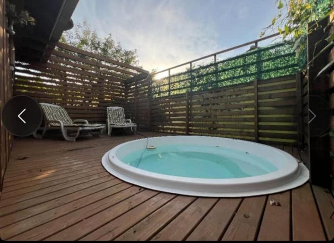 A wooden deck features a round jacuzzi surrounded by a privacy fence. Two lounge chairs are positioned nearby, inviting relaxation. The scene is softened by natural light filtering through the surrounding greenery.
