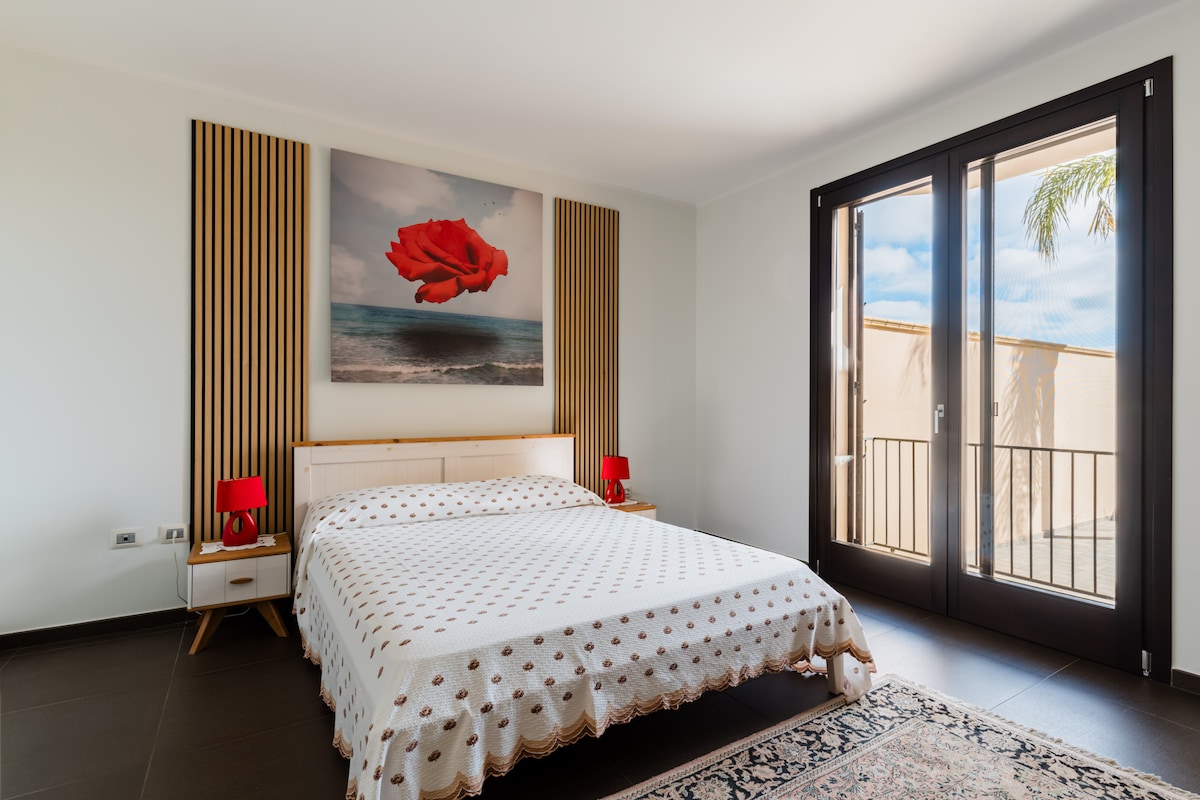 The bedroom features a spacious bed with a patterned coverlet. Two red bedside lamps provide accent lighting, and a large artwork of a rose hangs above the headboard. Floor-to-ceiling windows allow natural light to fill the room, with access to a balcony visible.