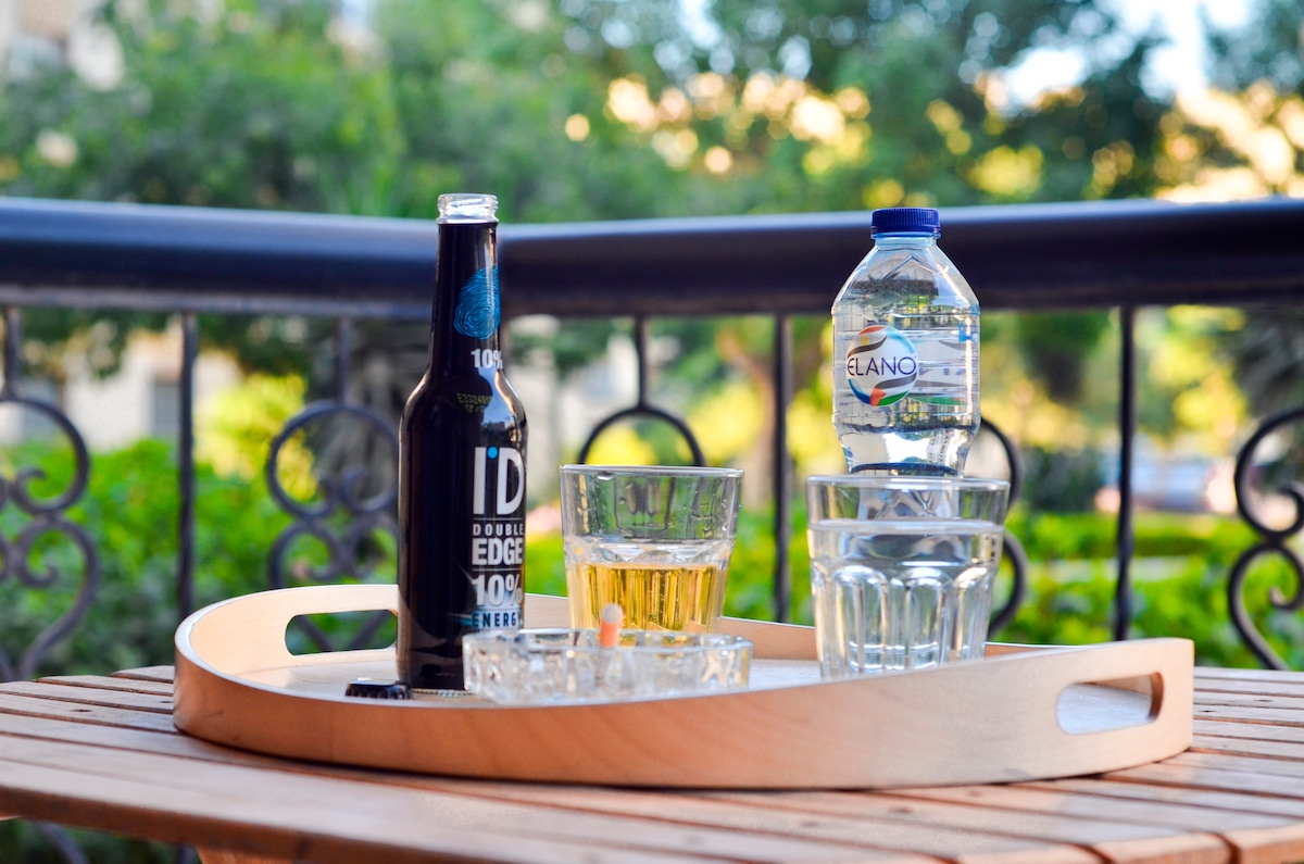 A wooden tray is displayed on a table, featuring a glass filled with a light-colored drink, a beer bottle, and a bottle of water. Clear glasses and an ashtray are also on the tray, with greenery visible in the background, creating a calm setting.