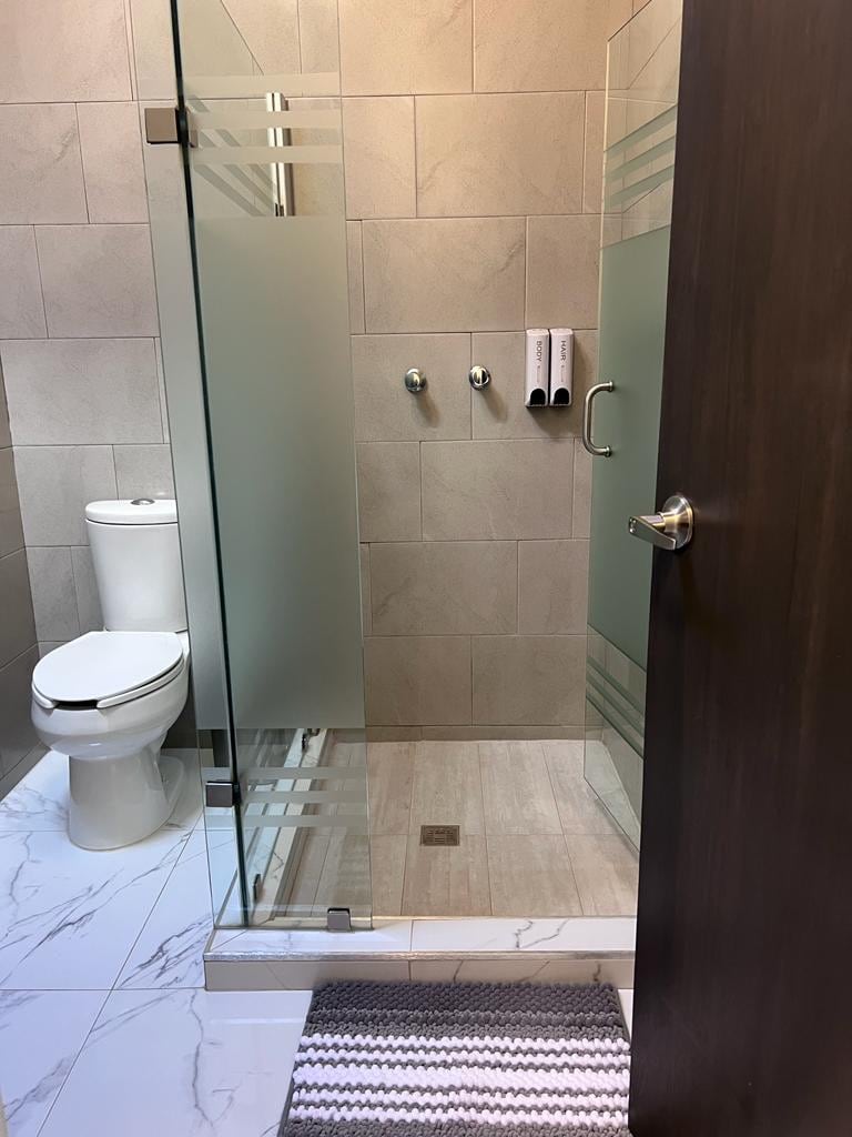 A modern bathroom is presented, featuring a spacious glass shower with sleek frosted panels. A white toilet is positioned adjacent to the shower. Light grey tiles cover the walls, while marbled flooring adds texture. A striped mat lies on the floor near the entrance.